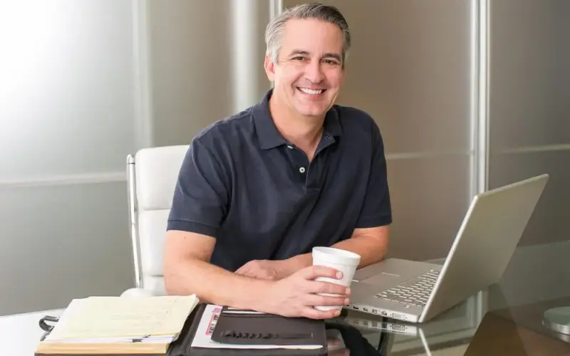 Imagem de um homem sorrindo, debruçado sob uma mesa com um notebook, pastas e um copo de café
