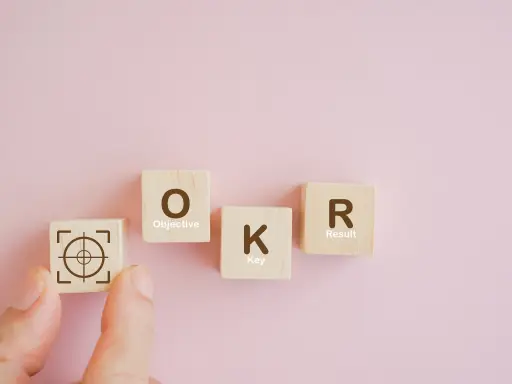 fundo rosa com 4 cubos de madeira onde o primeiro é um alvo e os outros formam a palavra okr